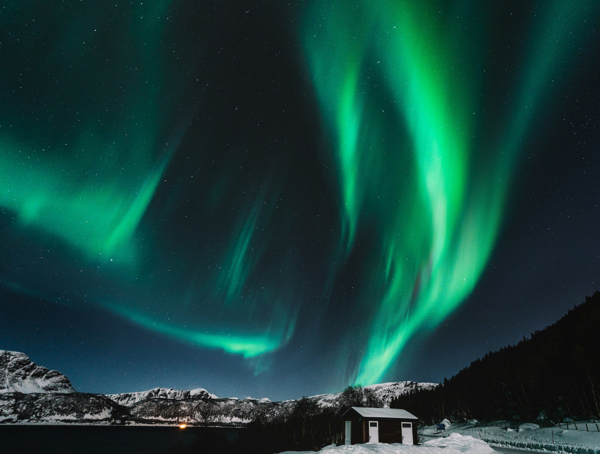 Grønt nordlys over fjellene og en liten hytte i snøen.